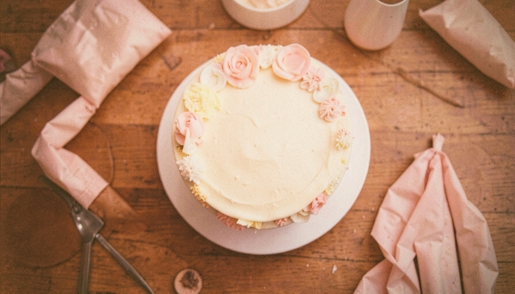 A softly lit overhead photograph of a half-decorated cake on a wooden table, with piping bags and fondant tools scattered nearby; warm cream and pale pink tones, visible film grain.