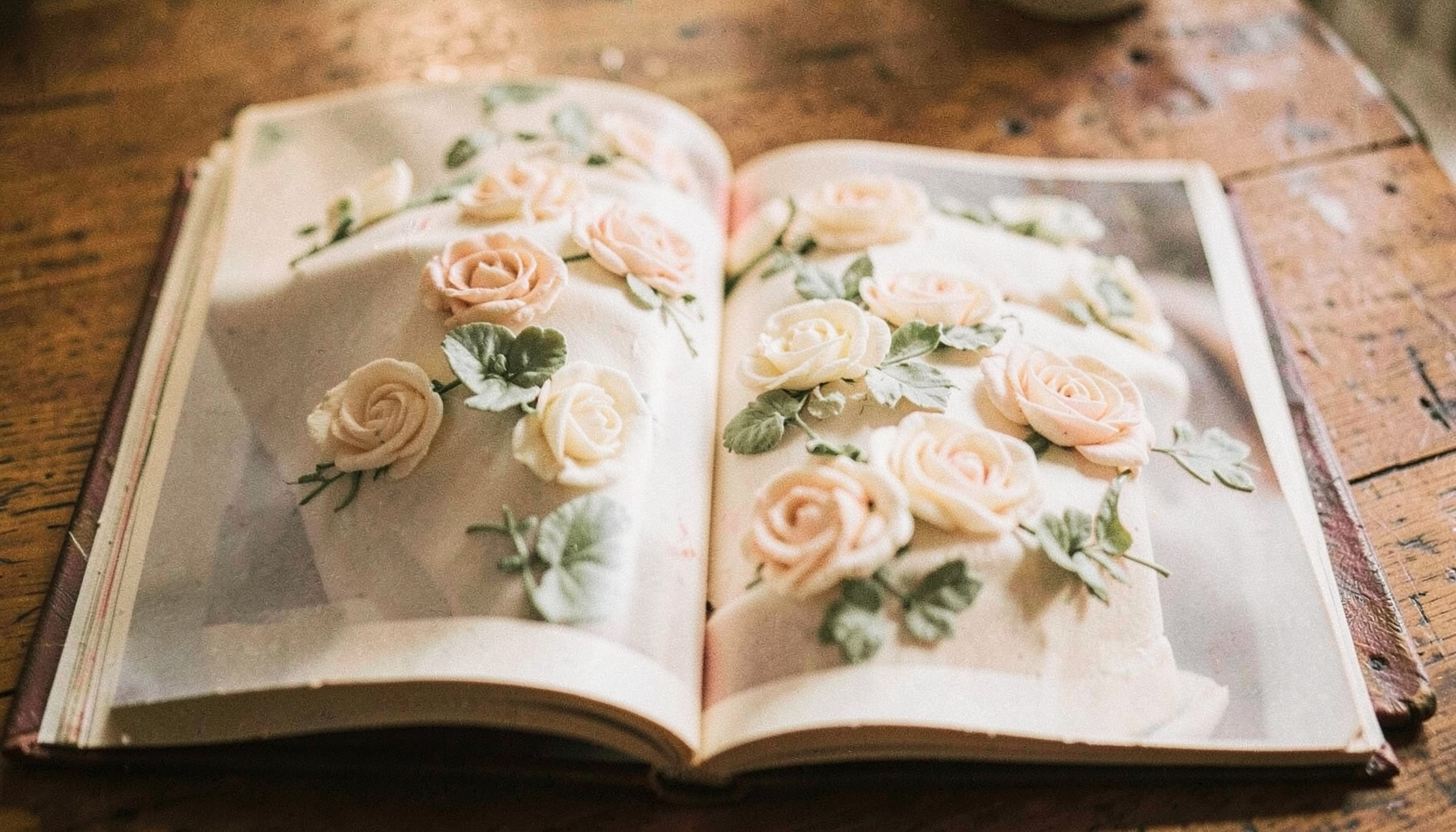 A well-worn instructional cake decorating book open on a wooden table, soft natural light falling across glossy pages showing piped buttercream flowers, warm cream and sage tones