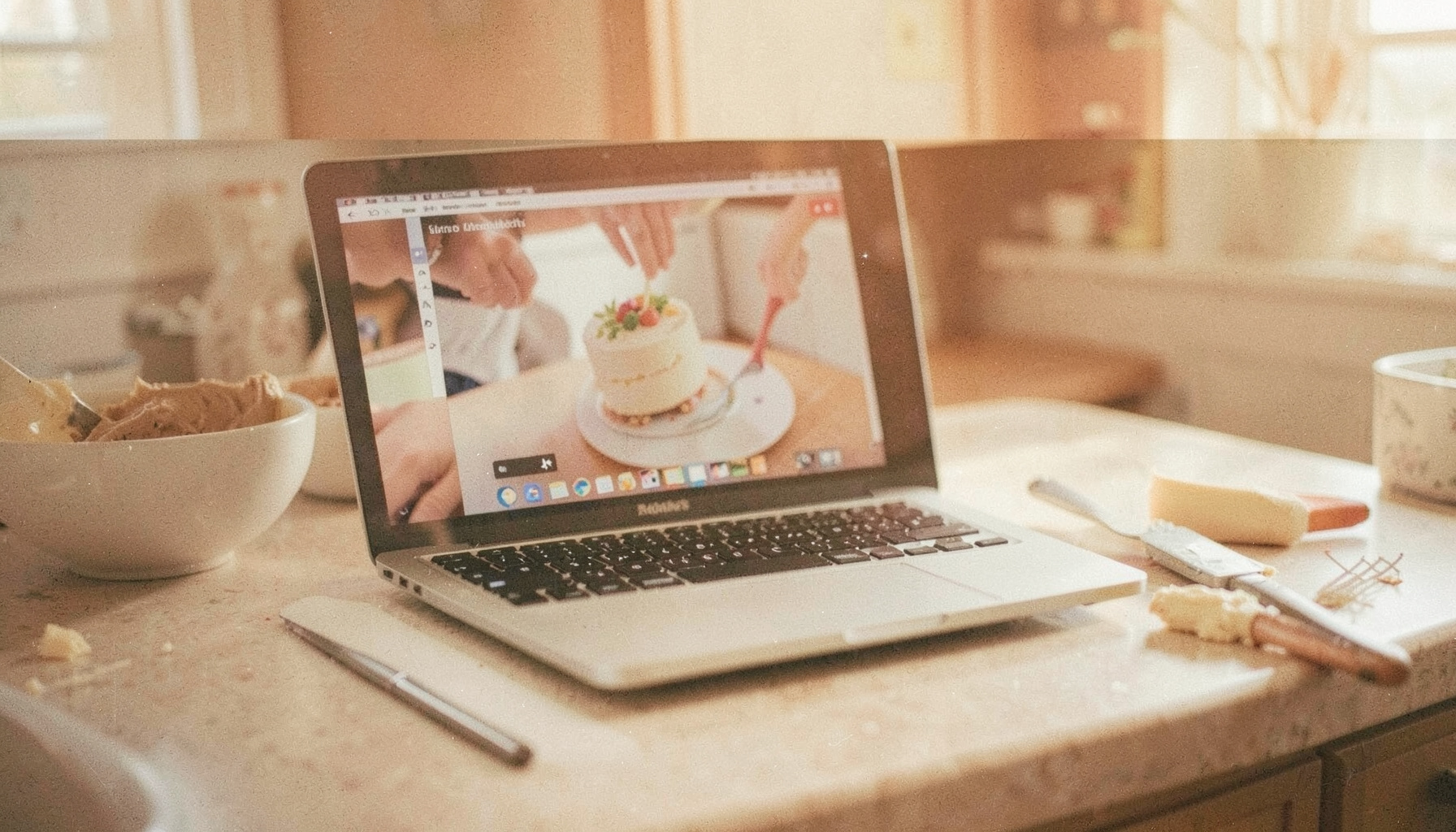 A laptop on a kitchen counter playing a cake decorating video, buttercream and spatula nearby, soft natural daylight, warm beige palette